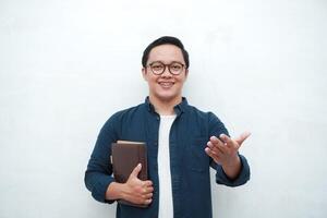 Young Asian man holding books smiling happy and positive while reading a book. Asian student pointing and showing welcoming gesture photo