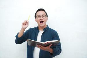 Happy young Asian man with glasses showing expression of gets genius idea. Asian college student showing happy and excited expression while holding book and pen photo