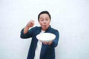Portrait of happy Asian man wearing a blue shirt while holding an empty plate with happy and excited expression. Young man showing and pointing empty mockup plate. Isolated with a white background. photo