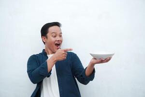 Portrait of happy Asian man wearing a blue shirt while holding an empty plate with happy and excited expression. Young man showing and pointing empty mockup plate. Isolated with a white background. photo