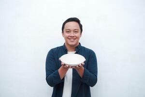 Portrait of happy Asian man wearing a blue shirt while holding an empty plate with happy and excited expression. Young man showing and pointing empty mockup plate. Isolated with a white background. photo
