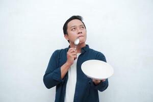 A portrait of happy Asian man wearing a blue shirt while thinking what to eat. Young man holding spoon and empty mockup plate. Isolated with a white background. photo
