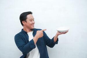 Portrait of happy Asian man wearing a blue shirt while holding an empty plate with happy and excited expression. Young man showing and pointing empty mockup plate. Isolated with a white background. photo
