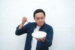 Portrait of happy Asian man wearing a blue shirt while holding an empty plate with happy and excited expression. Young man showing and pointing empty mockup plate. Isolated with a white background. photo
