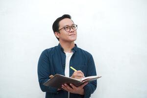 Happy young Asian man with glasses showing expression of gets genius idea. Asian college student showing happy and excited expression while holding book and pen photo