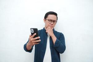 Young Asian man thinking and concentrating while holding books and handphone. Asian student thinking and looking for ideas while holding phone over white background photo