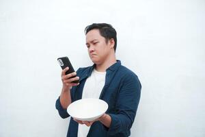 A portrait of happy Asian man wearing a blue shirt while thinking what to eat. Young man holding spoon and empty mockup plate. Isolated with a white background. photo