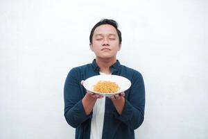 A portrait of happy Asian man wearing a blue shirt while holding a plate of fried noodles. Young man smiling and pointing to a plate of noodle. Isolated with a white background. photo