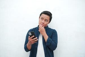 Young Asian man thinking and concentrating while holding books and handphone. Asian student thinking and looking for ideas while holding phone over white background photo