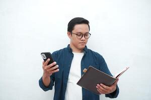 Asian college student holding book and blank smartphone screen with happy and excited expression. Young Asian student wearing blue shirt smiling and showing white mobile phone screen photo