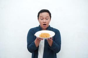 A portrait of happy Asian man wearing a blue shirt while holding a plate of fried noodles. Young man smiling and pointing to a plate of noodle. Isolated with a white background. photo