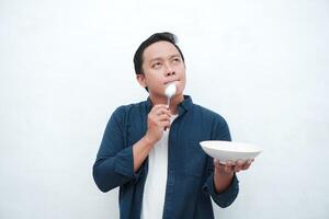 A portrait of happy Asian man wearing a blue shirt while thinking what to eat. Young man holding spoon and empty mockup plate. Isolated with a white background. photo