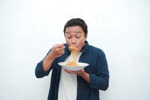A portrait of happy Asian man wearing a blue shirt while holding a plate of fried noodles. Young man smiling and pointing to a plate of noodle. Isolated with a white background. photo