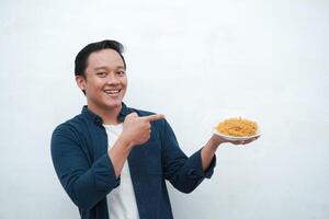 A portrait of happy Asian man wearing a blue shirt while holding a plate of fried noodles. Young man smiling and pointing to a plate of noodle. Isolated with a white background. photo