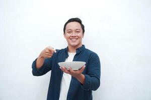 Portrait of happy Asian man wearing a blue shirt while holding an empty plate with happy and excited expression. Young man showing and pointing empty mockup plate. Isolated with a white background. photo