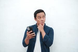 Young Asian man thinking and concentrating while holding books and handphone. Asian student thinking and looking for ideas while holding phone over white background photo