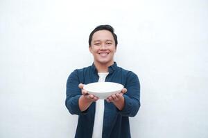 Portrait of happy Asian man wearing a blue shirt while holding an empty plate with happy and excited expression. Young man showing and pointing empty mockup plate. Isolated with a white background. photo
