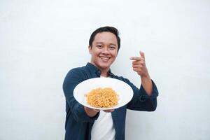 A portrait of happy Asian man wearing a blue shirt while holding a plate of fried noodles. Young man smiling and pointing to a plate of noodle. Isolated with a white background. photo