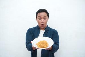 A portrait of happy Asian man wearing a blue shirt while holding a plate of fried noodles. Young man smiling and pointing to a plate of noodle. Isolated with a white background. photo