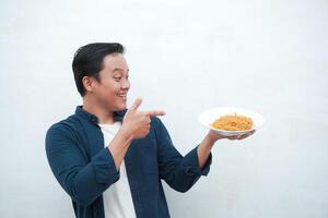 A portrait of happy Asian man wearing a blue shirt while holding a plate of fried noodles. Young man smiling and pointing to a plate of noodle. Isolated with a white background. photo