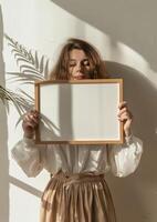 Woman Holding Blank Frame Mockup Against White Wall with Sunlit Shadows in Minimalistic Setting. photo