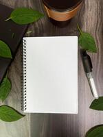 Still life photography blank notebook on wooden table with a cup of coffee, plant, leaf on table photo