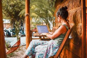 Close up young caucasian hipster woman sit use computer read news media articles, write travel blog or browse internet search or work online while traveling in southeast asia tropical islands photo