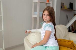 Child with a cast on a broken wrist or arm smiling and having fun on a couch. Positive attitude, recovery and kid concept. photo