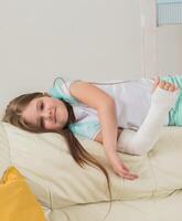 Child with a cast on a broken wrist or arm smiling and having fun on a couch. Positive attitude, recovery and kid concept. photo