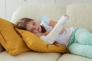 Sad child with a cast on a broken wrist or arm lying on a couch. Recovery and disease concept. photo