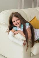 Child with a cast on a broken wrist or arm smiling and having fun on a couch. Positive attitude, recovery and kid concept. photo
