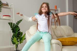 Child with a cast on a broken wrist or arm smiling and having fun on a couch. Positive attitude, recovery and kid concept. photo