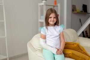 Child with a cast on a broken wrist or arm smiling and having fun on a couch. Positive attitude, recovery and kid concept. photo
