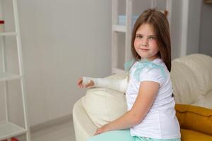 Child with a cast on a broken wrist or arm smiling and having fun on a couch. Positive attitude, recovery and kid concept. photo