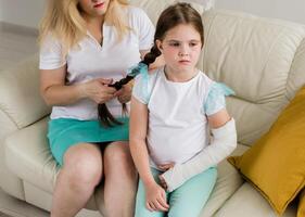 Adorable preschooler girl with a broken arm orthopedic cast at home on sofa with mother. Broken limb and broken arm in a cast in kids photo