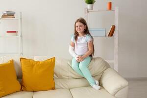 Child with a cast on a broken wrist or arm smiling and having fun on a couch. Positive attitude, recovery and kid concept. photo