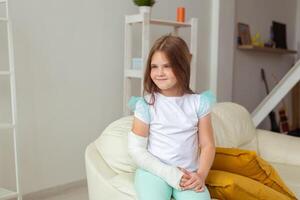Child with a cast on a broken wrist or arm smiling and having fun on a couch. Positive attitude, recovery and kid concept. photo