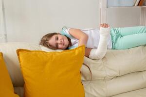 Child with a cast on a broken wrist or arm smiling and having fun on a couch. Positive attitude, recovery and kid concept. photo