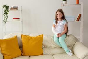 Child with a cast on a broken wrist or arm smiling and having fun on a couch. Positive attitude, recovery and kid concept. photo