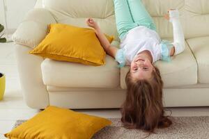 Child with a cast on a broken wrist or arm smiling and having fun on a couch. Positive attitude, recovery and kid concept. photo