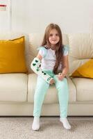 Child with a cast on a broken wrist or arm smiling and having fun on a couch. Positive attitude, recovery and kid concept. photo