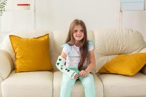 Child with a cast on a broken wrist or arm smiling and having fun on a couch. Positive attitude, recovery and kid concept. photo