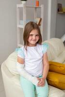 Child with a cast on a broken wrist or arm smiling and having fun on a couch. Positive attitude, recovery and kid concept. photo