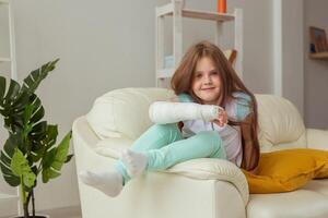 Child with a cast on a broken wrist or arm smiling and having fun on a couch. Positive attitude, recovery and kid concept. photo