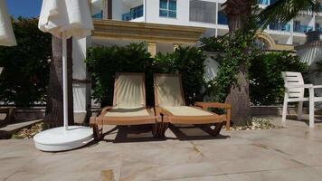 Two sun loungers bask in the shadow of palm branches, creating a serene scene against the backdrop of a building. The interplay of light and shadow on the sun loungers adds to the tranquil atmosphere. video