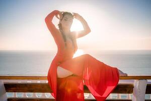 Closeup portrait of sensual young brunette woman in red dress, happily dancing outdoors isolated on blurry sea background with natural bokeh in soft warm sunset backlight. Selective focus. photo