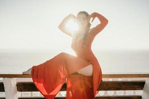 Closeup portrait of sensual young brunette woman in red dress, happily dancing outdoors isolated on blurry sea background with natural bokeh in soft warm sunset backlight. Selective focus. photo