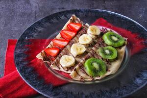 Colorful Fruit Crepe with Chocolate opped with fresh strawberries, bananas, and kiwi slices, with rich chocolate sauce served in plate isolated on table side view of indulgent dessert photo