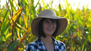 Happy smiling female farmer looks into camera standing near corn field. Portrait of adult beautiful agronomist in straw hat with maize meadow at background. Agricultural business concept. Close up photo
