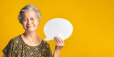 Concept of question mark speech bubble. An elderly Asian woman with short gray hair holding a blank paper and looking at the camera with a smile while standing on a yellow background photo